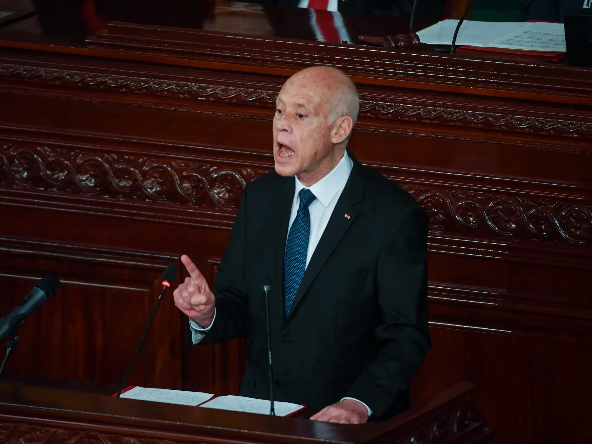 A bald man in a dark suit speaks at a wooden lectern, raising one finger beside microphones.