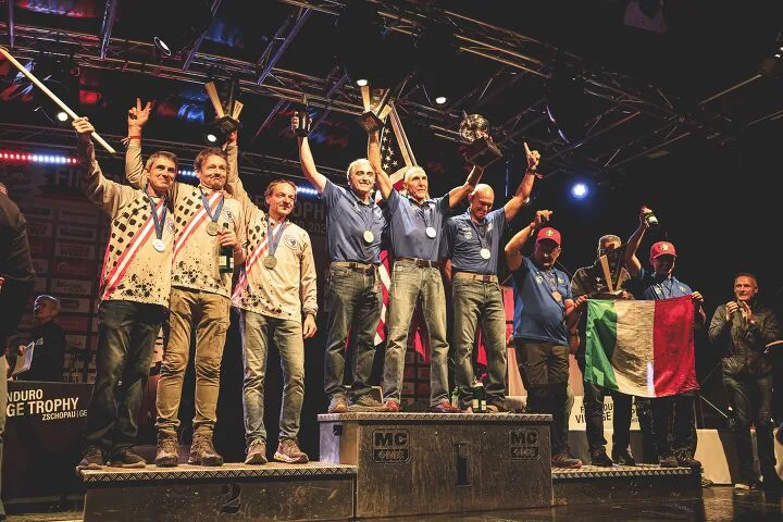 Men stand on an awards podium holding trophies, with a team displaying an Italian flag.