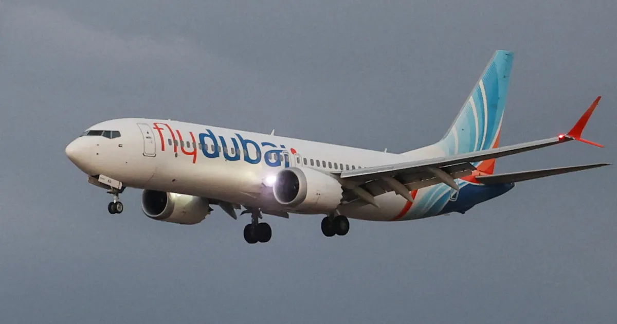 A Flydubai passenger jet flies with its landing gear lowered against a gray sky.
