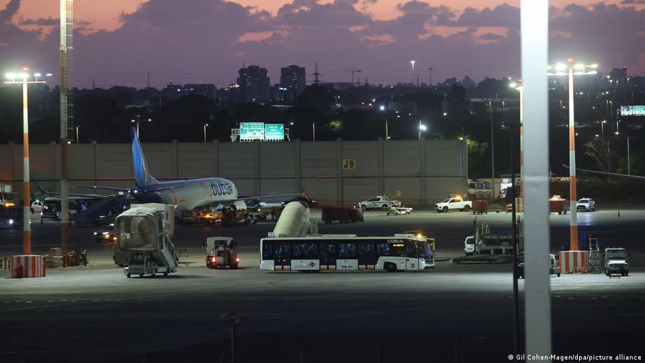 Airliner, airport shuttle bus, and ground service vehicles on a floodlit airport apron at dusk.