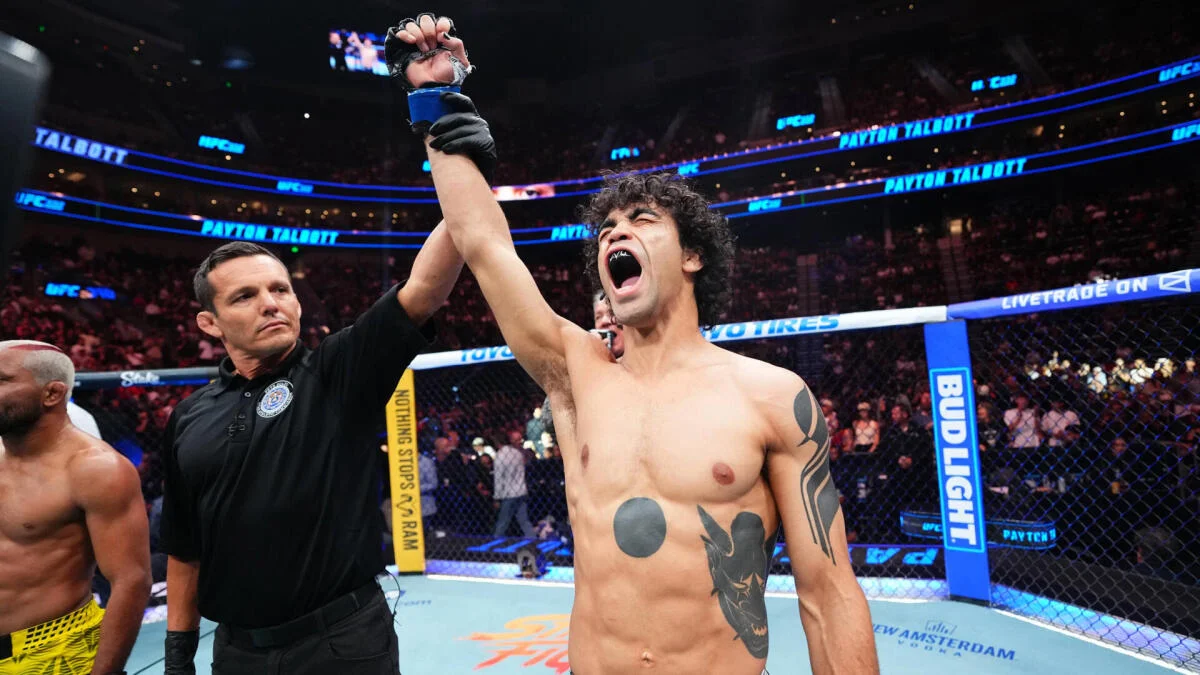 A referee raises a shirtless fighter’s gloved hand inside a crowded UFC octagon.