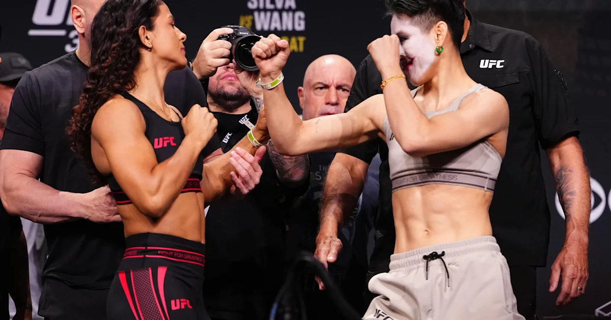 Two female fighters face off with raised fists at a UFC weigh-in stage, surrounded by officials and photographers.