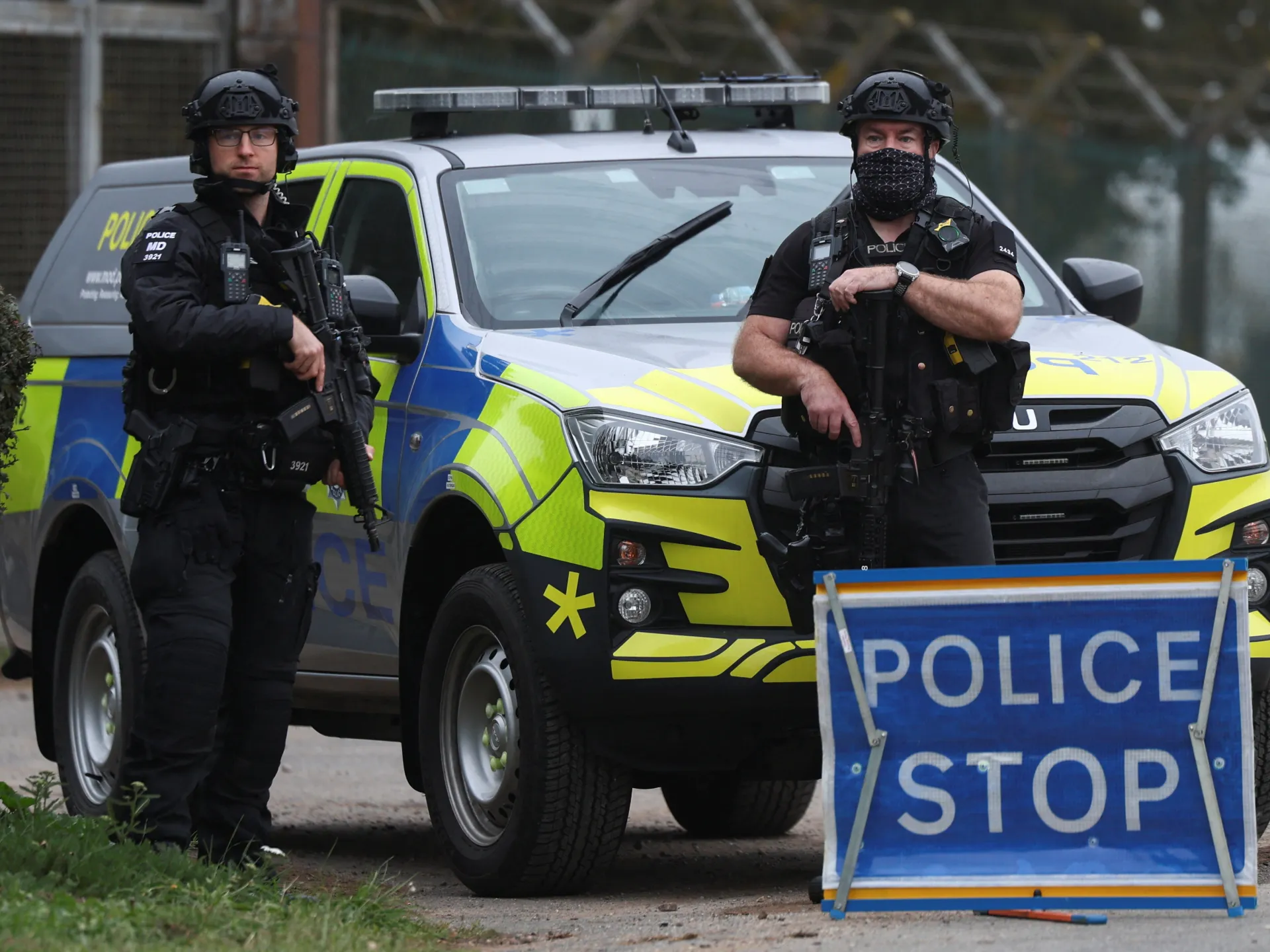 Two armed officers stand beside a marked police vehicle and a police stop sign.