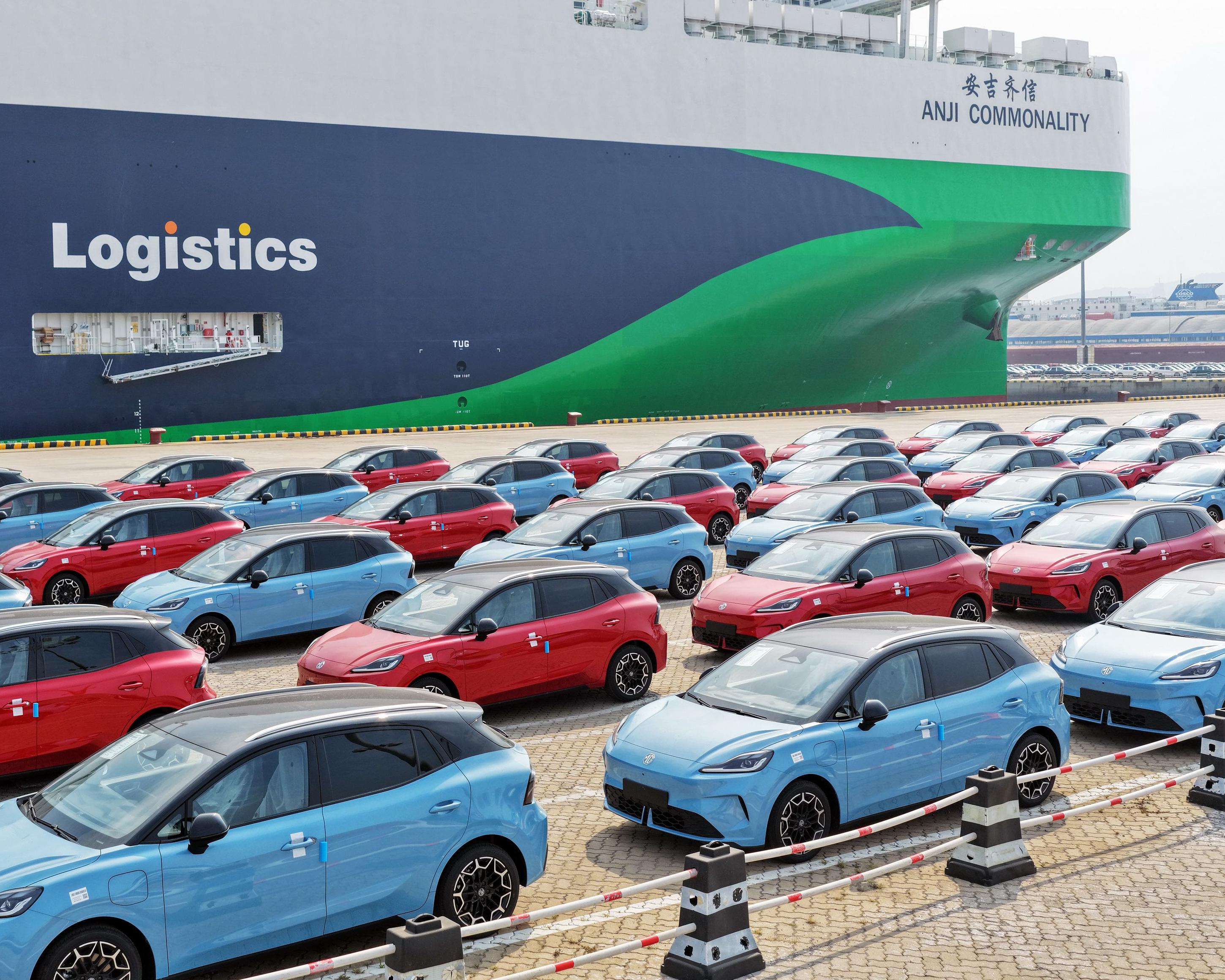 Rows of red and blue cars are parked beside a large green and navy cargo ship.