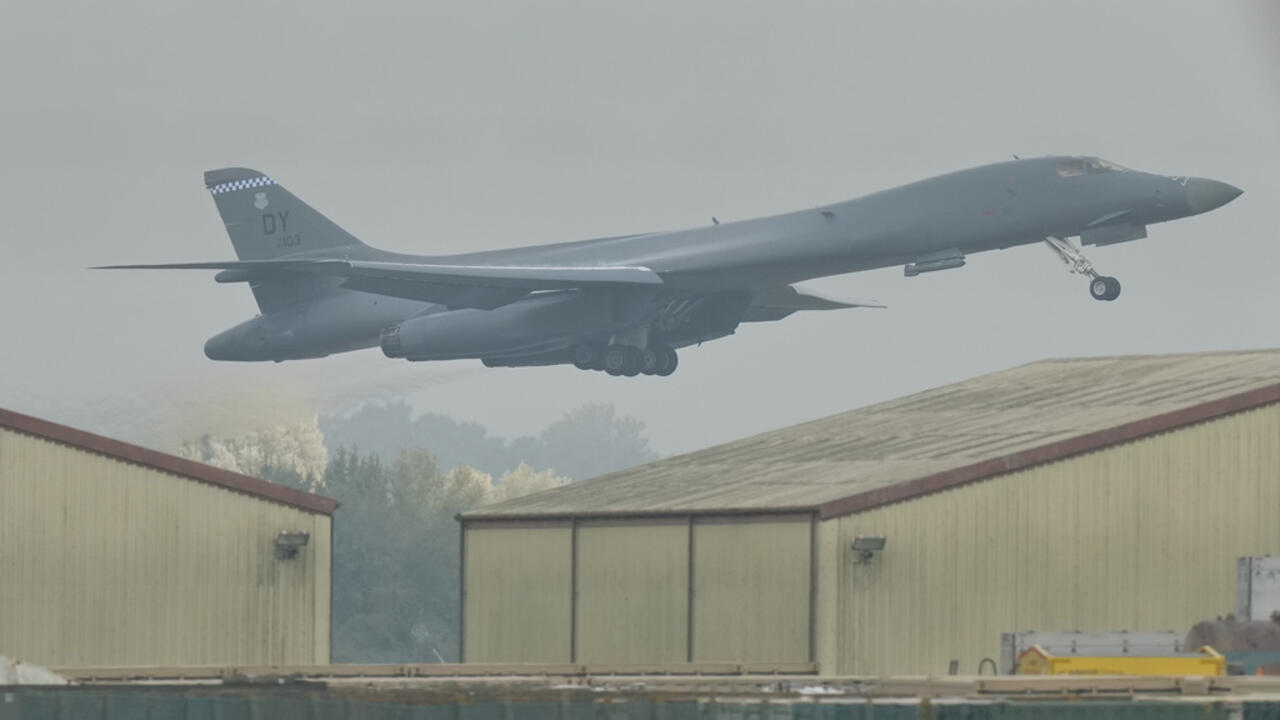 A military bomber takes off above hangars, with its landing gear still extended.