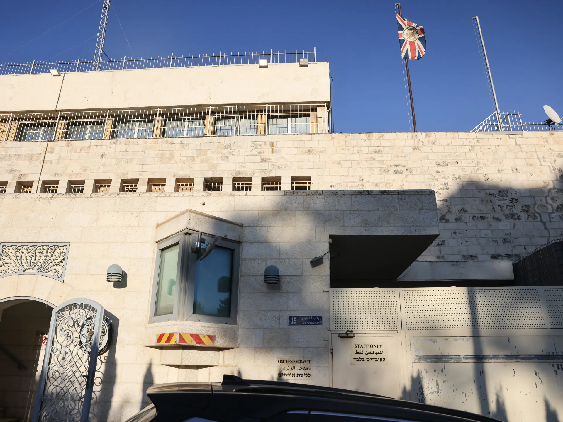 Sunlit stone building with barred windows, an ornate gate, a staff entrance, and a Union Jack flag.