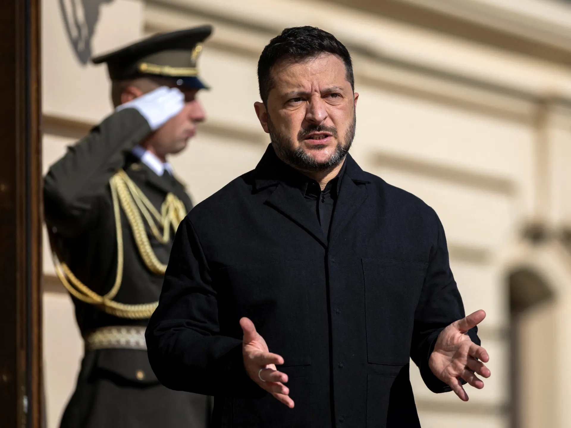 A man in a black coat gestures while speaking, with a uniformed guard saluting behind him.