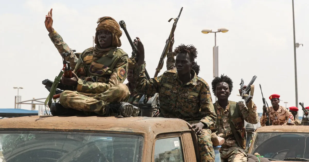 Armed men in military-style clothing ride on vehicles, with one man raising two fingers.
