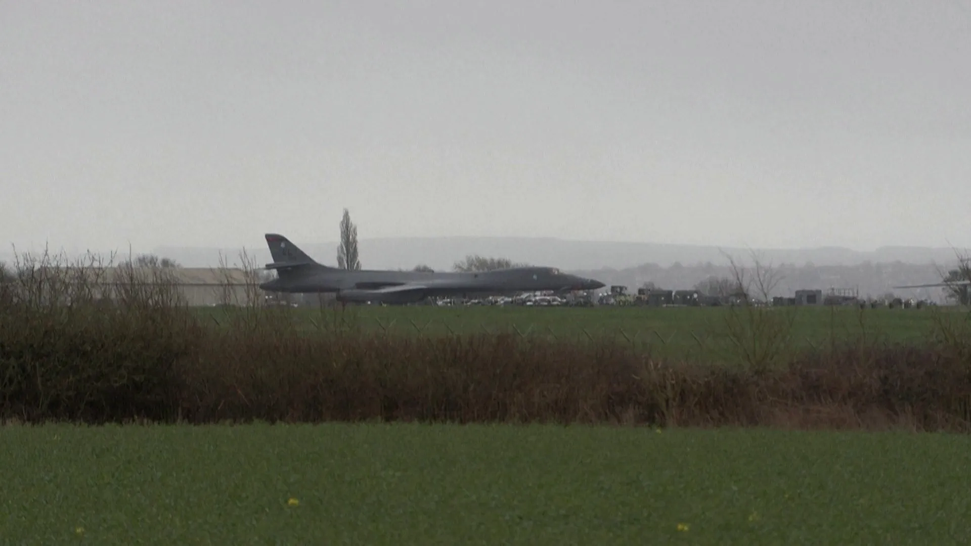 A dark bomber sits on an airfield beyond a green field under an overcast sky.