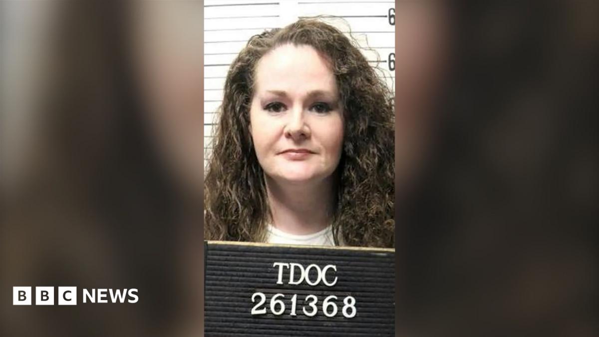 A woman with long curly hair holds a prison booking board in front of a height chart.