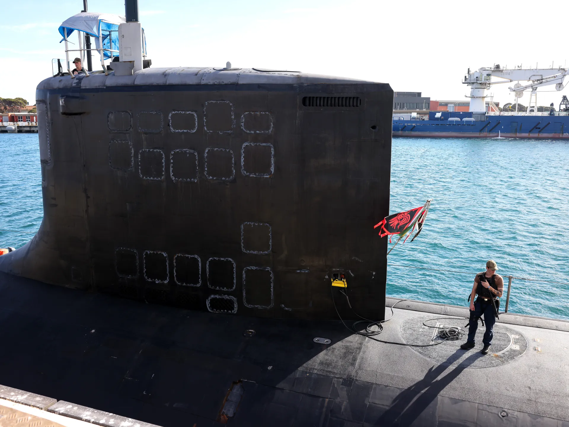 A dark submarine beside a dock, with a crew member standing near cables and a red flag.