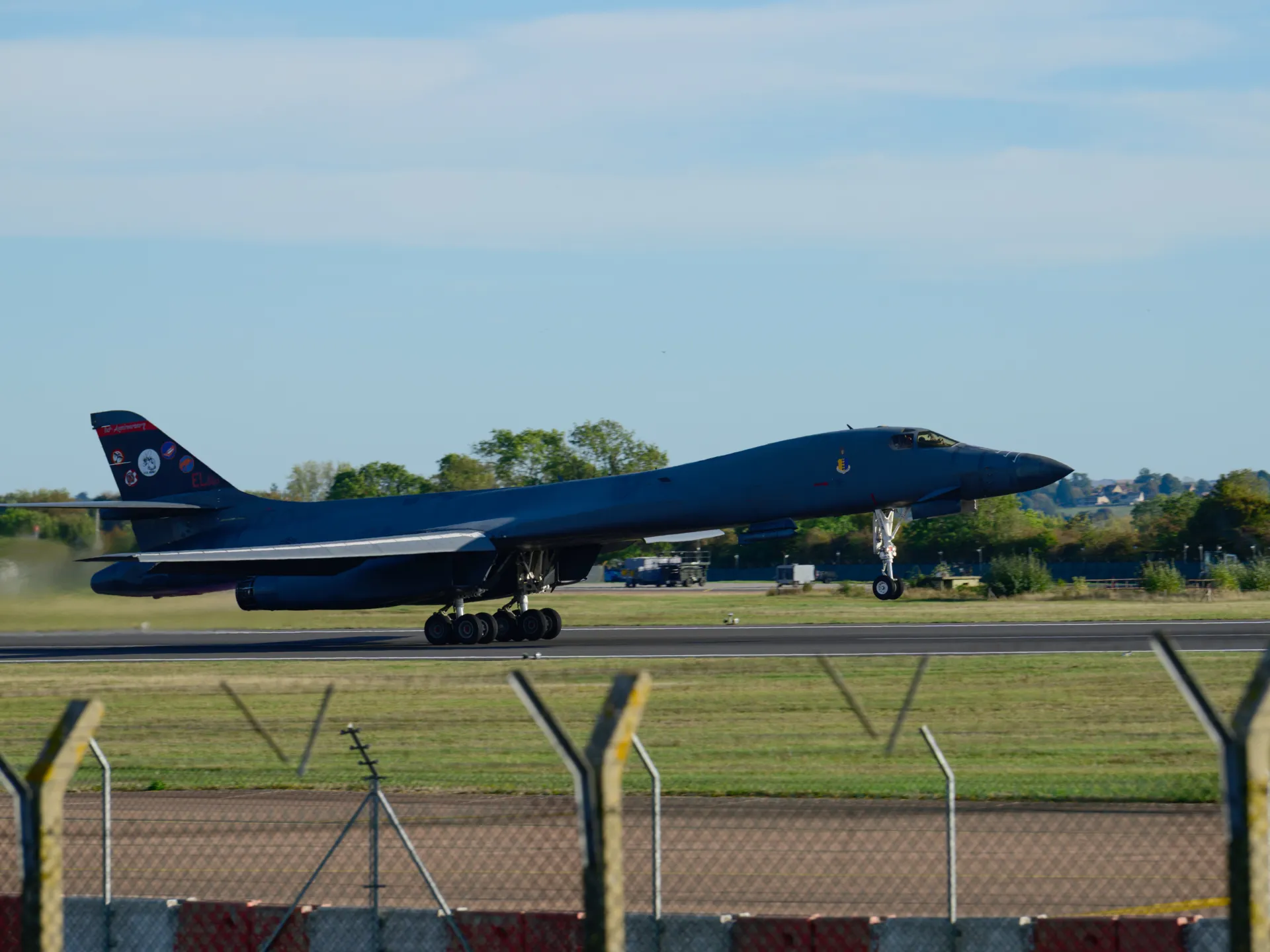 A dark military bomber rolls along a runway behind a foreground security fence under a clear sky.