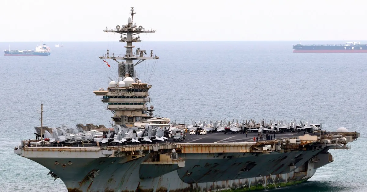 Aircraft carrier crowded with military aircraft sails past several cargo ships at sea.