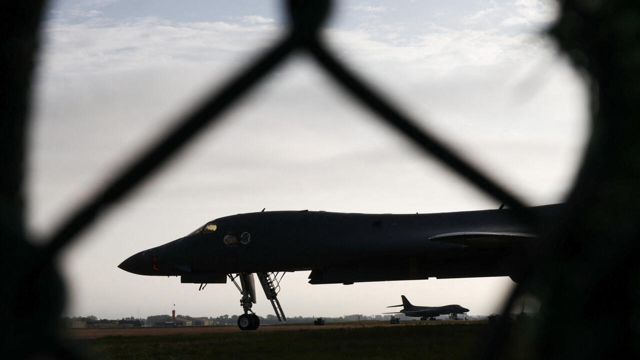 A silhouetted bomber sits on an airfield behind a dark, out-of-focus fence, with another aircraft in the distance.