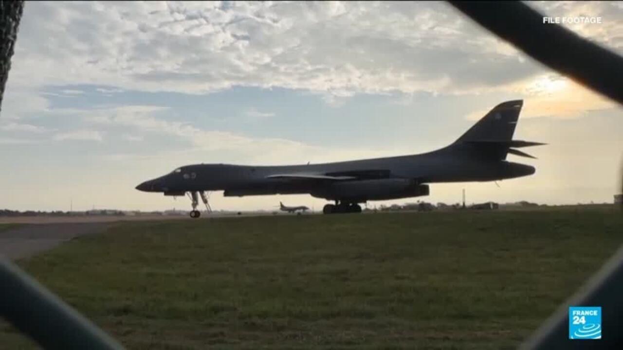 A dark bomber aircraft sits on a runway at dusk, viewed through a fence.