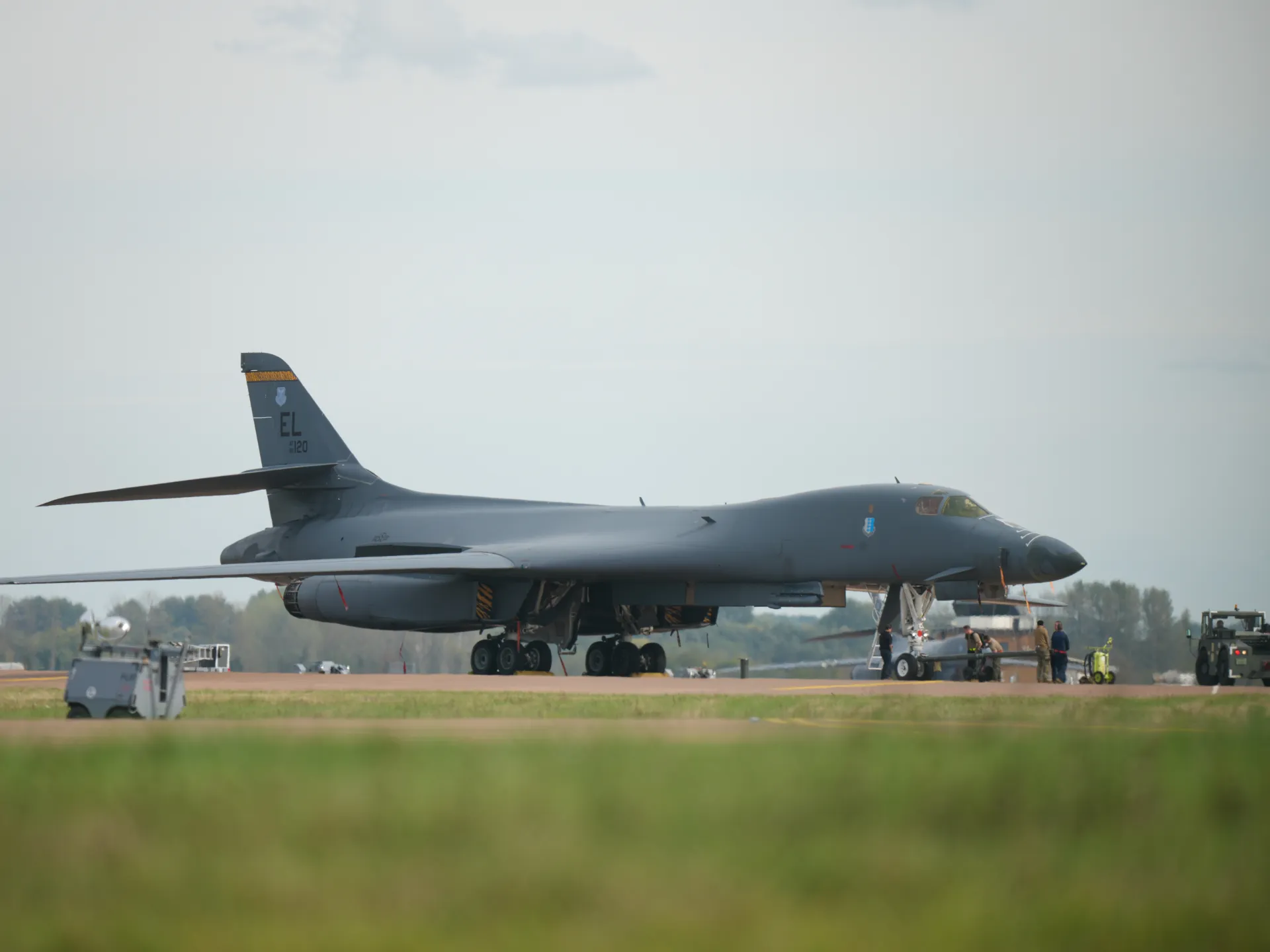 A gray bomber sits on an airfield as ground crew and service vehicles work nearby.