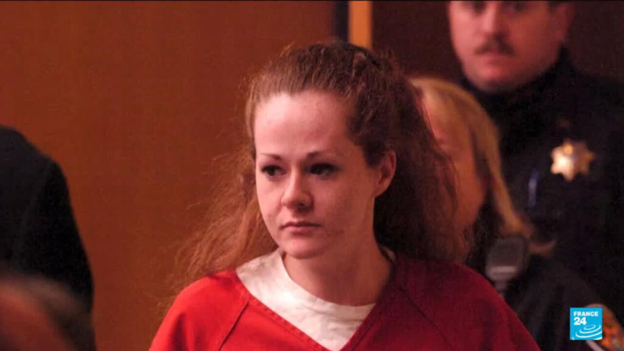 A woman in a red top sits among people indoors, looking to her left.
