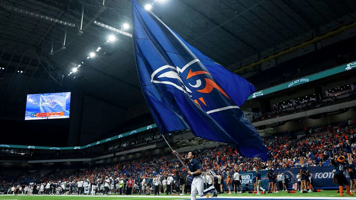 A person runs across the field waving a large blue and orange flag before a packed football crowd.