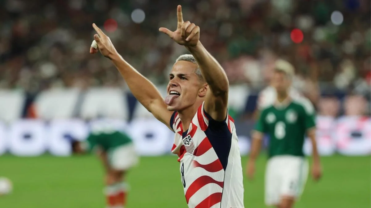 A U.S. player celebrates with both arms raised as a Mexico player bends forward in the background.