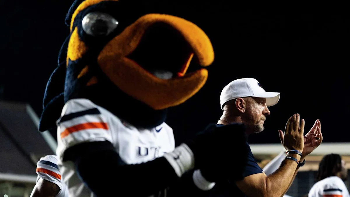 A bird mascot stands beside a man in a white cap clapping on the sideline at night.