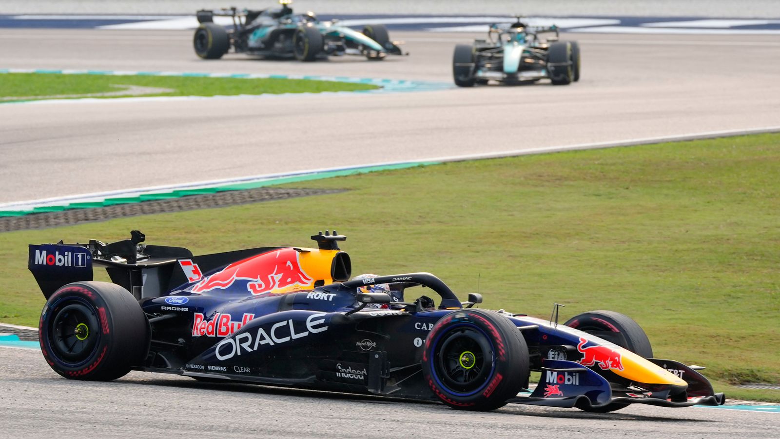 A Red Bull Formula One car leads two others around a circuit, with grass beside the track.