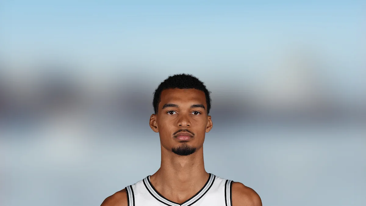 A basketball player in a white jersey faces the camera against a blurred indoor background.