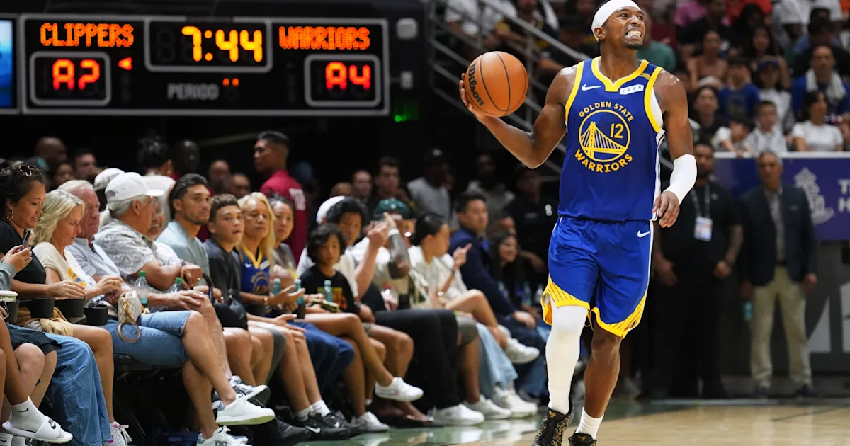 A Warriors player holds a basketball near courtside spectators, with the Clippers and Warriors scoreboard visible.