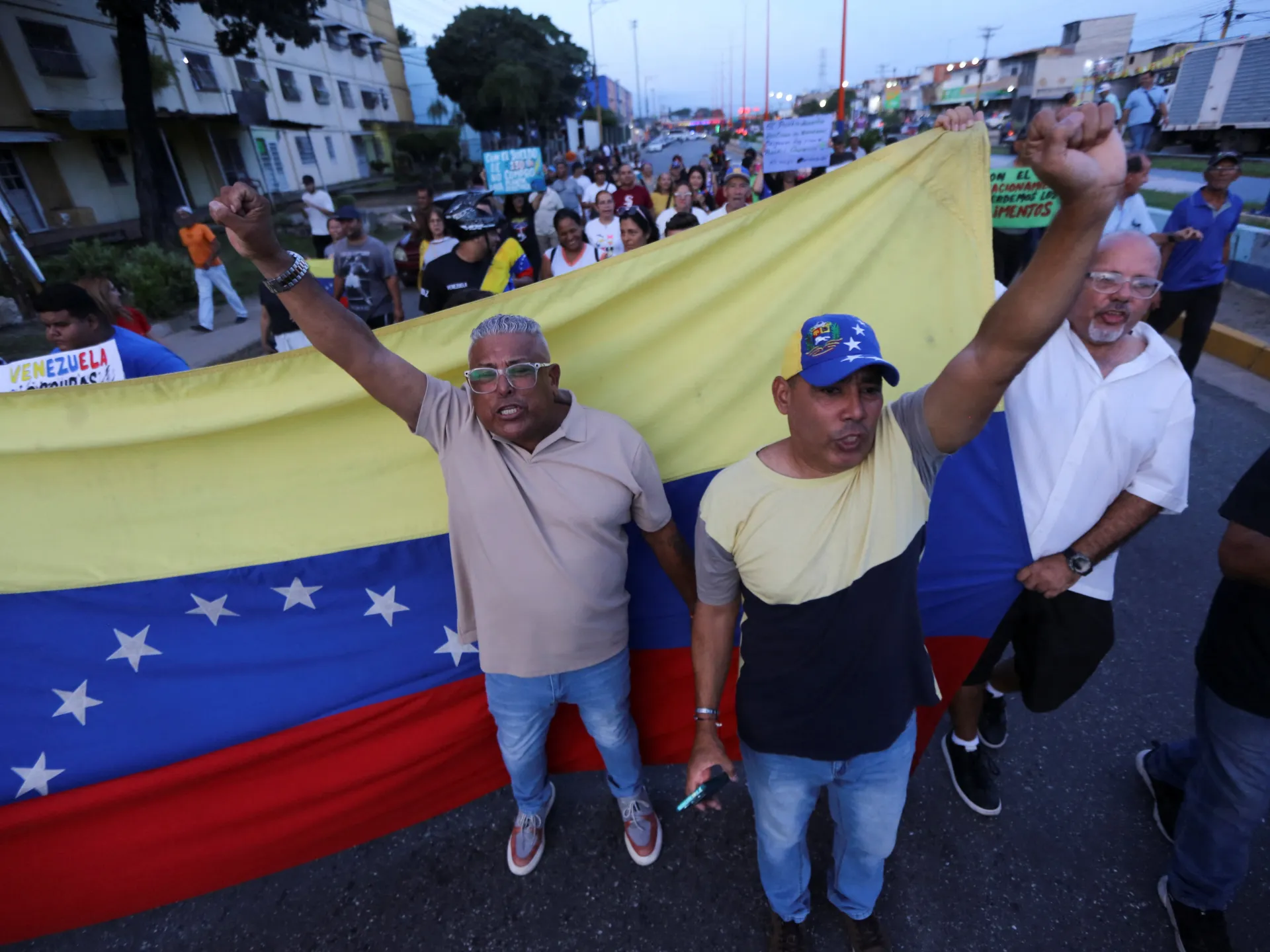 Demonstrators carry a large Venezuelan flag, with several men raising their fists at the front.
