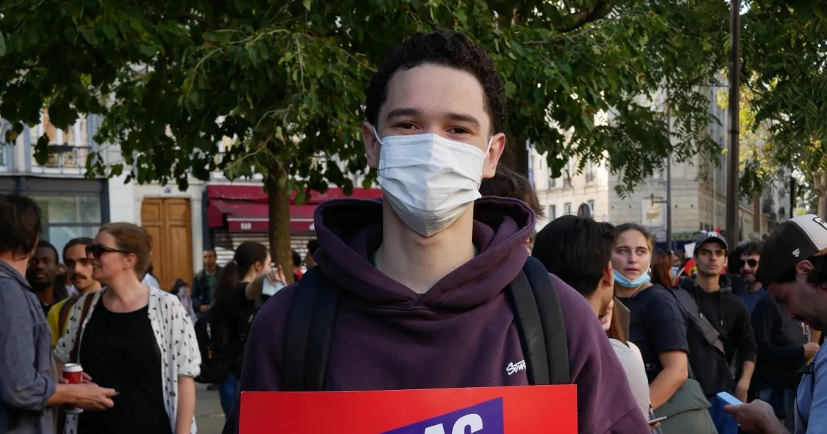 A masked young man in a purple hoodie stands among a crowd, holding a partially visible sign.