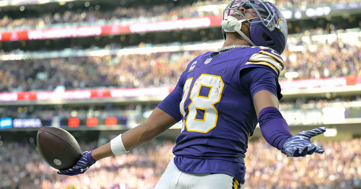 Football player in a purple number 18 uniform holds a ball in a stadium.