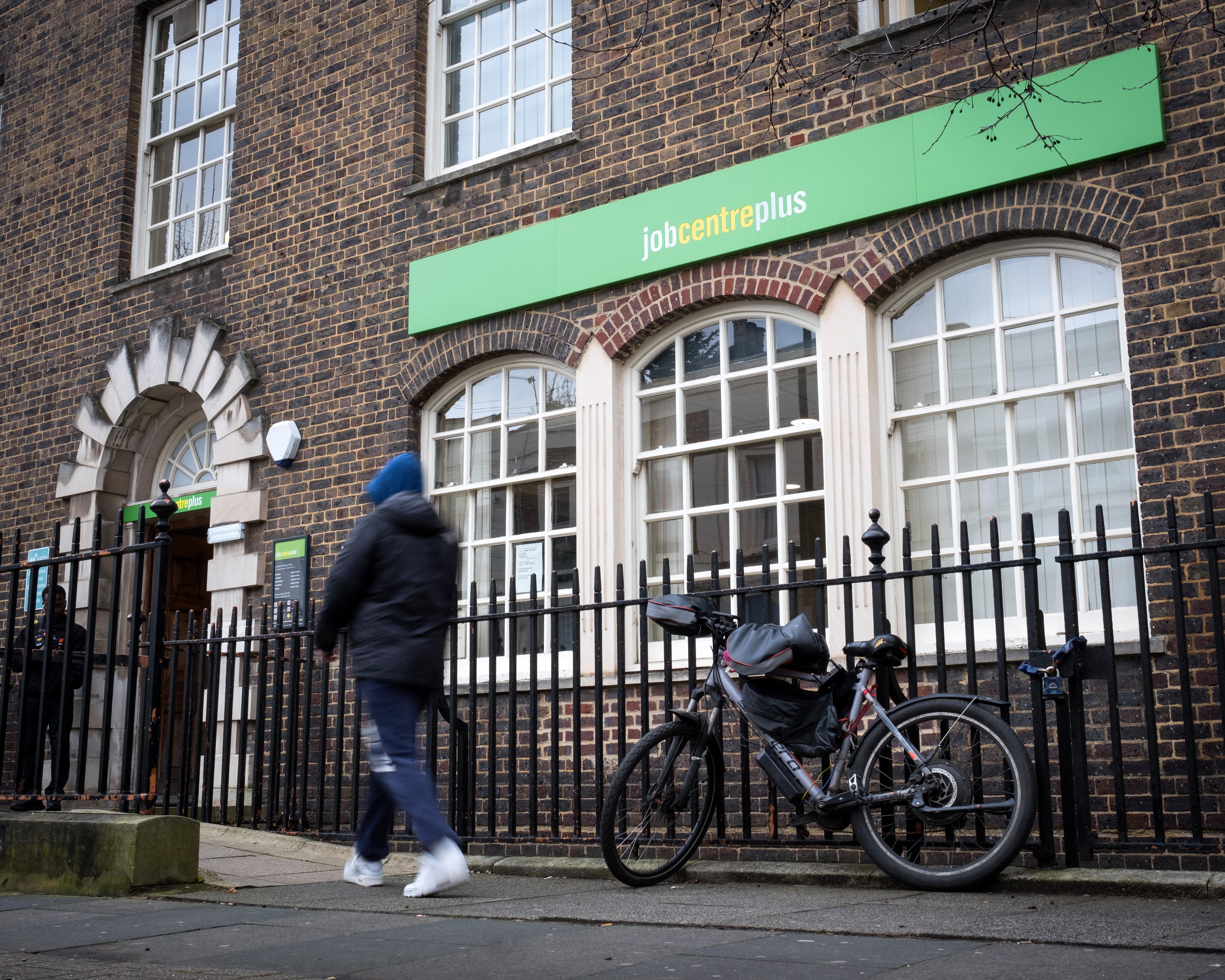 A blurred pedestrian passes a brick Jobcentre Plus building, with bicycles parked behind a metal fence.