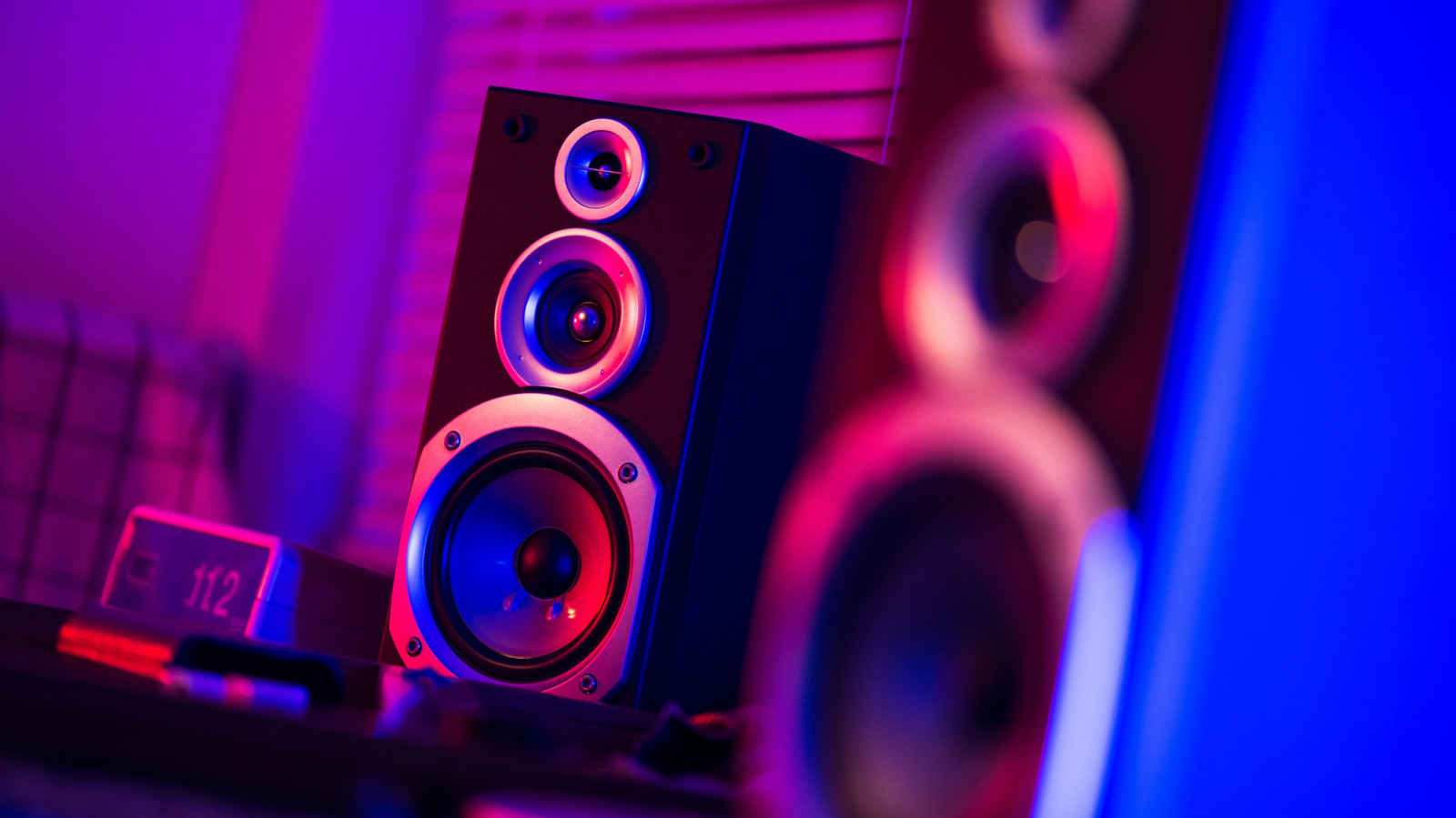 A three-driver speaker glows under magenta and blue lighting beside a blurred speaker cabinet.