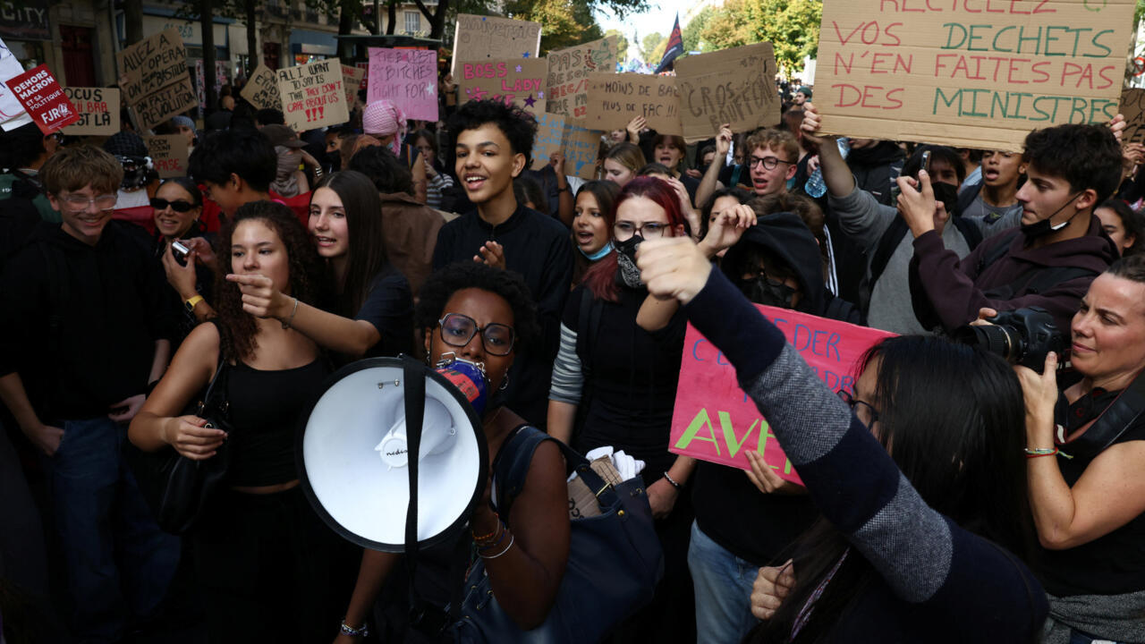 Students and other protesters crowd a street, holding handmade signs as a woman speaks through a megaphone.