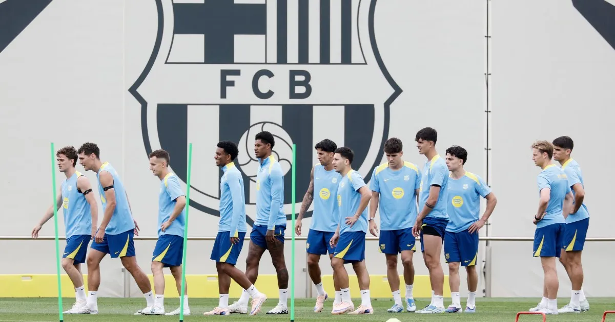 Football players in blue training gear walk across a pitch in front of a large club crest.