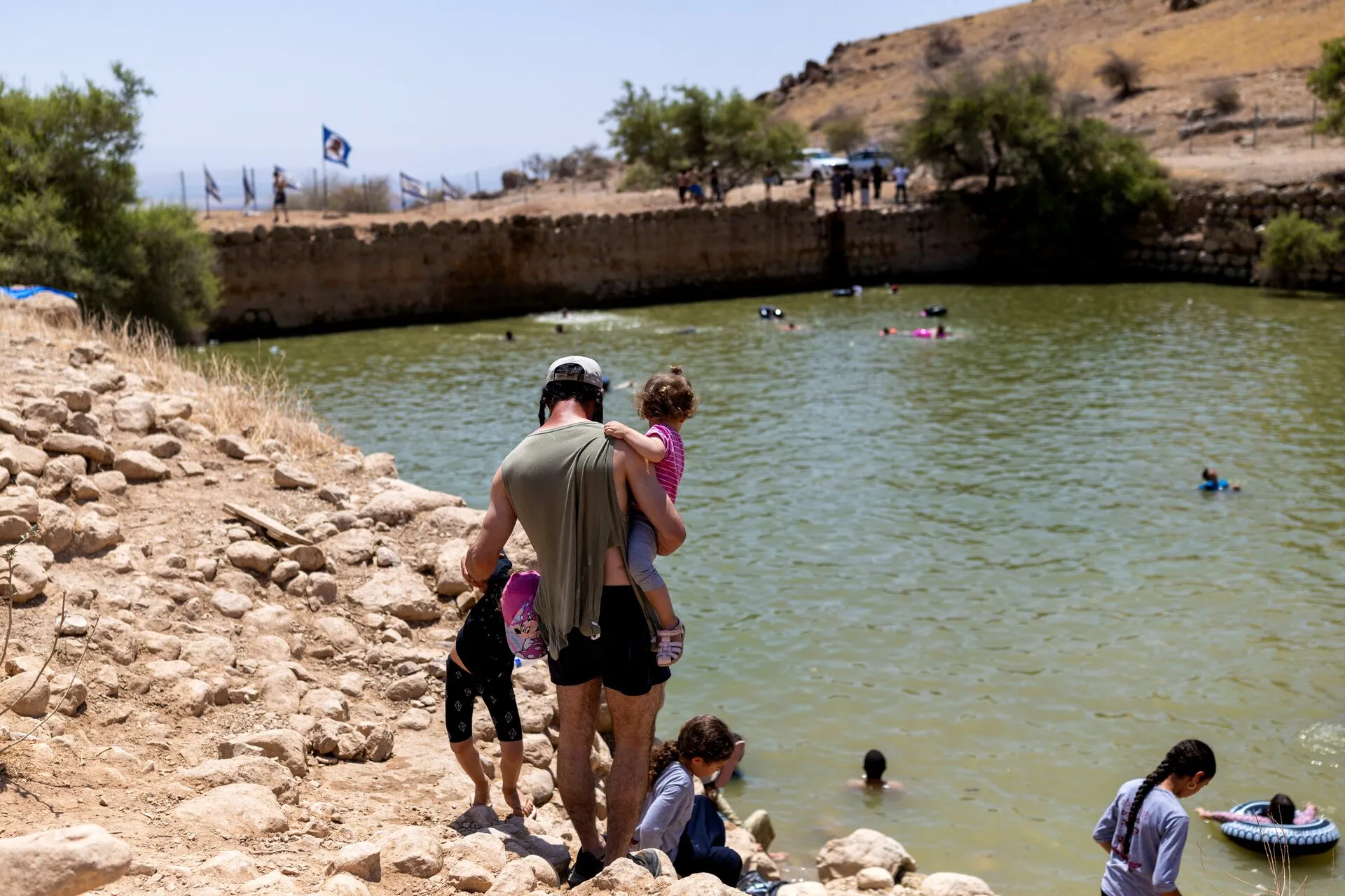 A man carries a young child beside a stone-edged pool where people are swimming.