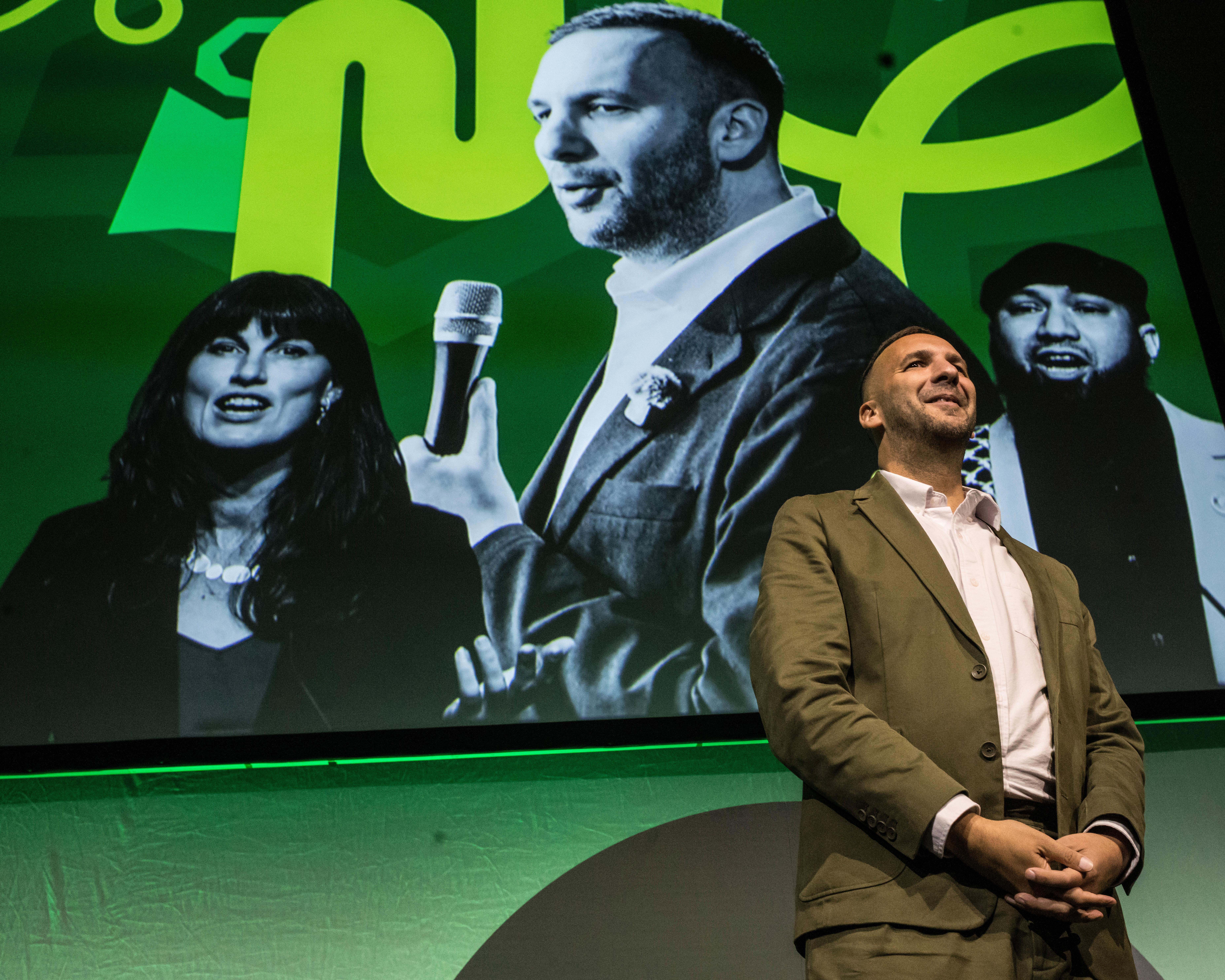 A seated man onstage beneath a large green screen showing three black-and-white portraits.