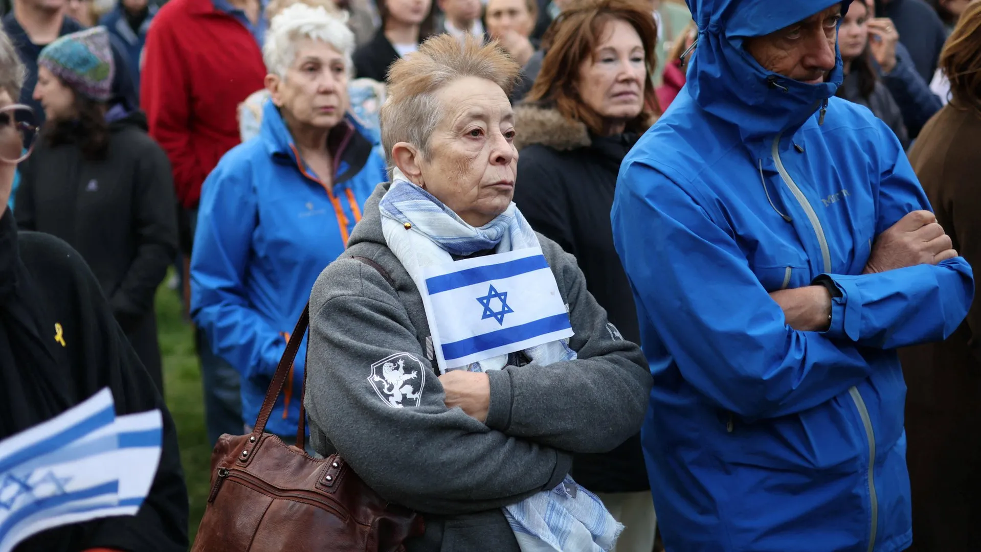 An older woman wearing an Israeli flag scarf stands among a crowd, arms crossed.