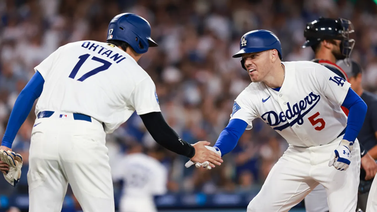 Two Dodgers players in white uniforms exchange a handshake on the field during a game.