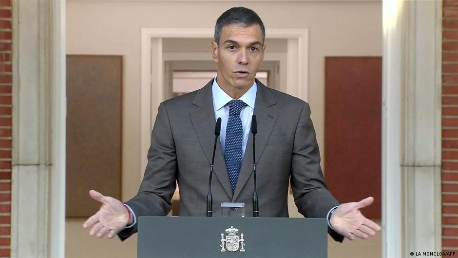 A suited man speaks at a lectern with two microphones, gesturing with both hands.