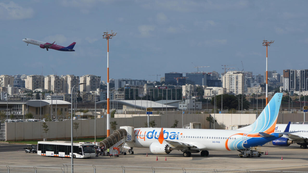 A Flydubai plane is parked at an airport gate as another airliner climbs above the city.