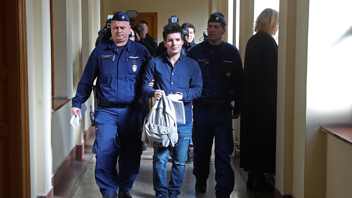 A man carrying a gray bag walks between uniformed officers in a courthouse corridor.