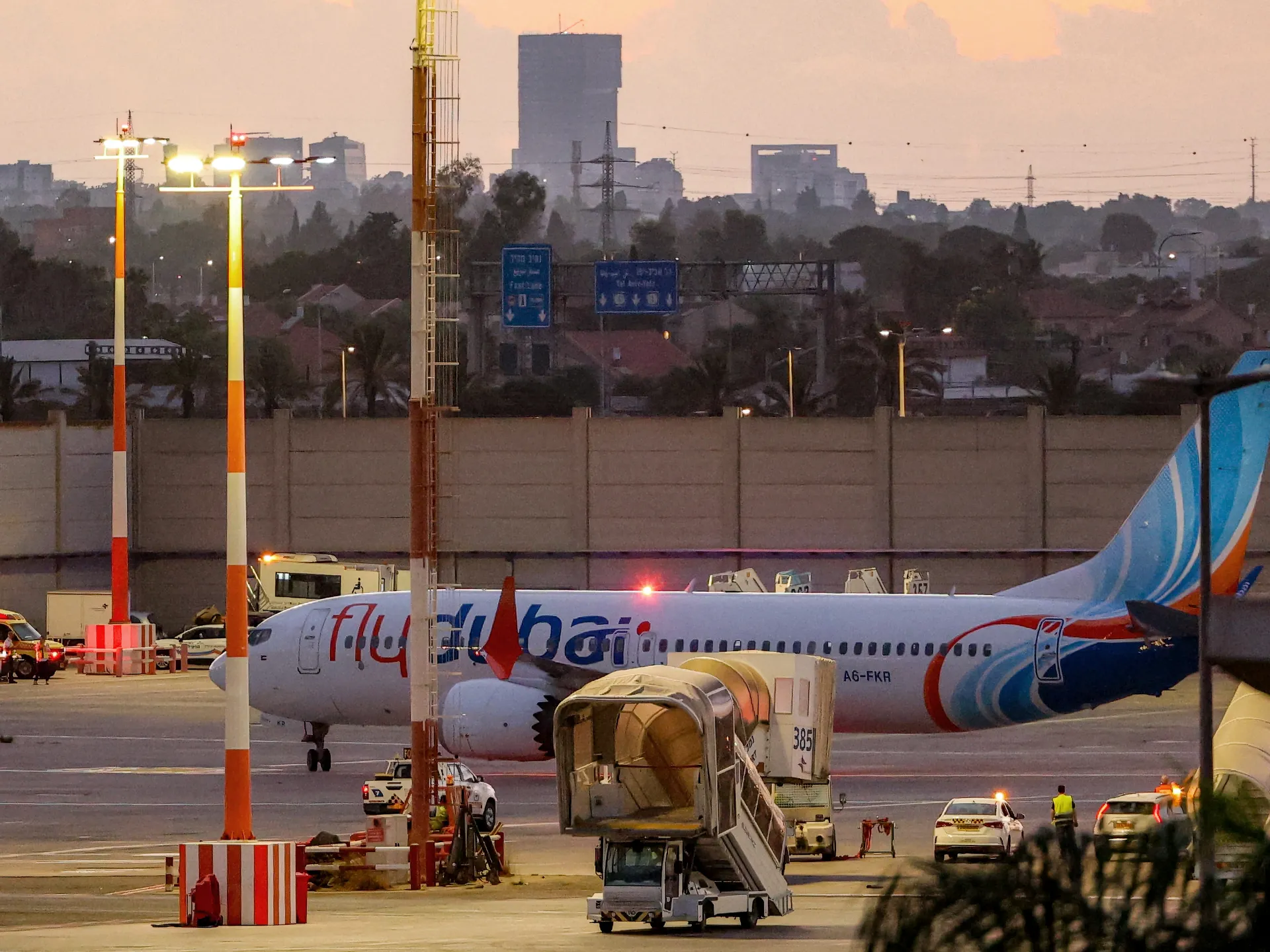 A Flydubai passenger jet sits on an airport apron at dusk among ground vehicles and boarding equipment.