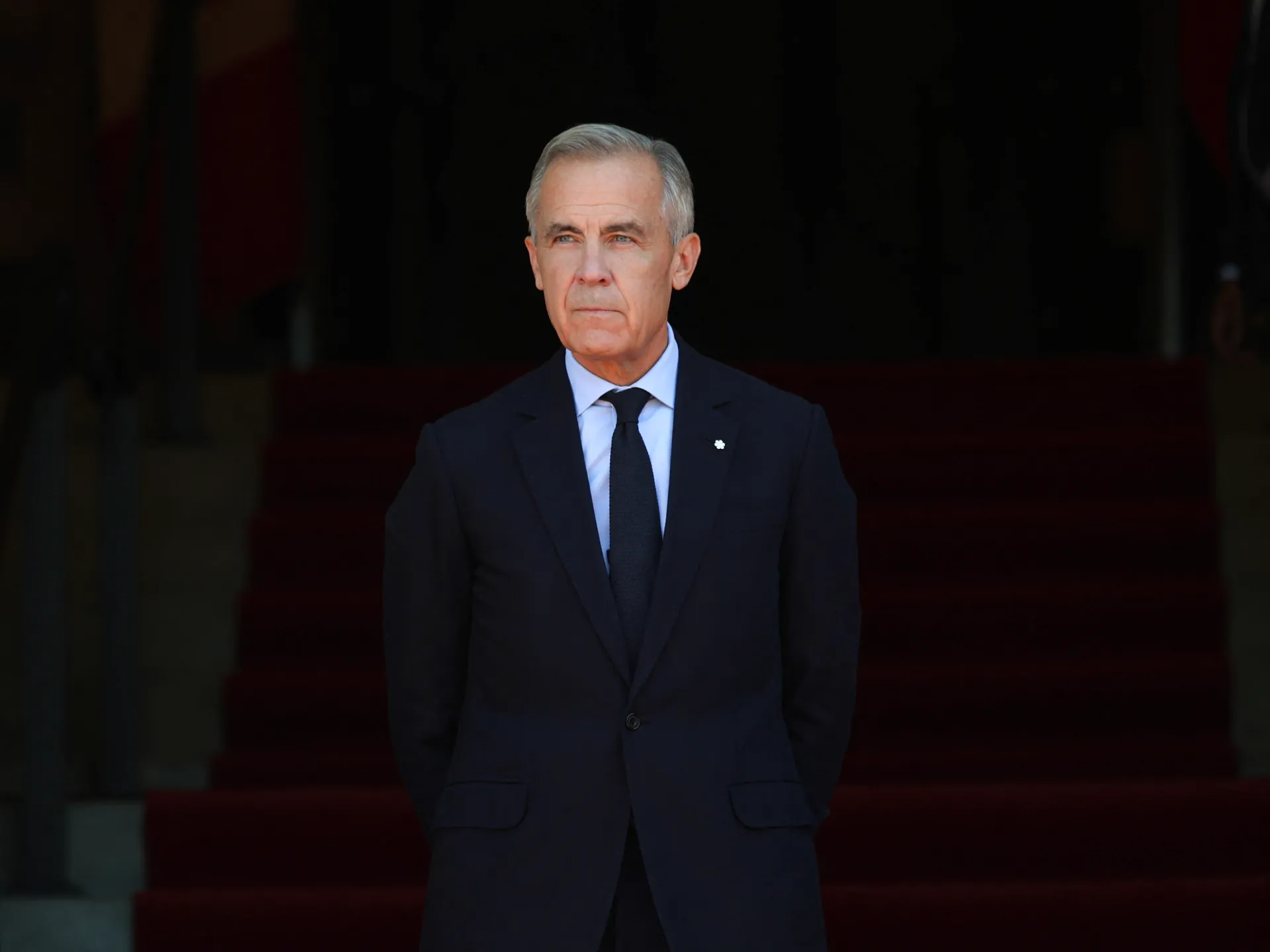 A gray-haired man in a dark suit stands with his hands behind his back against a dark red backdrop.