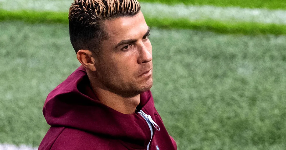 A footballer in a burgundy hoodie looks to the side against a blurred green field.