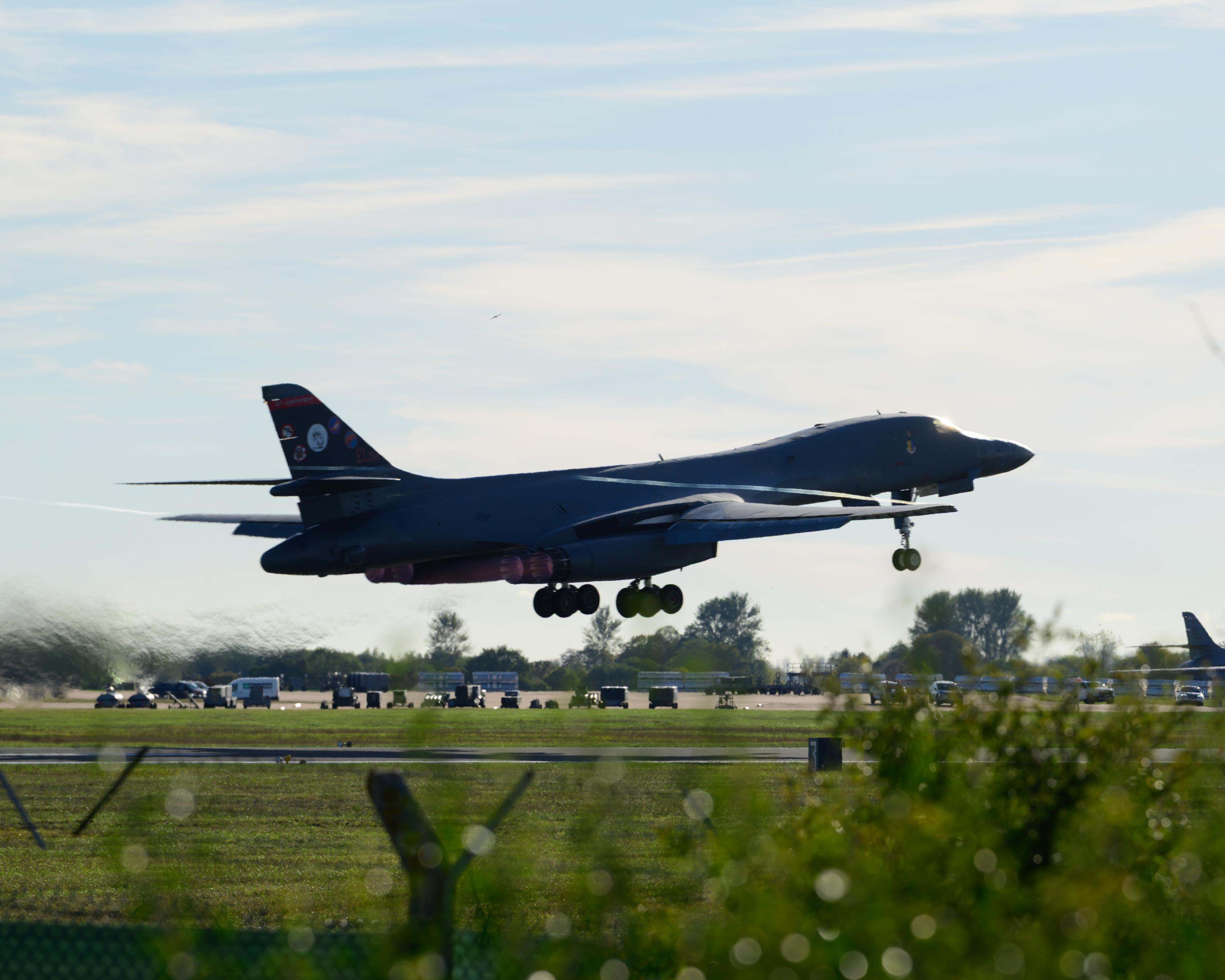 A dark military bomber descends toward a runway with landing gear extended, above a grassy airfield.