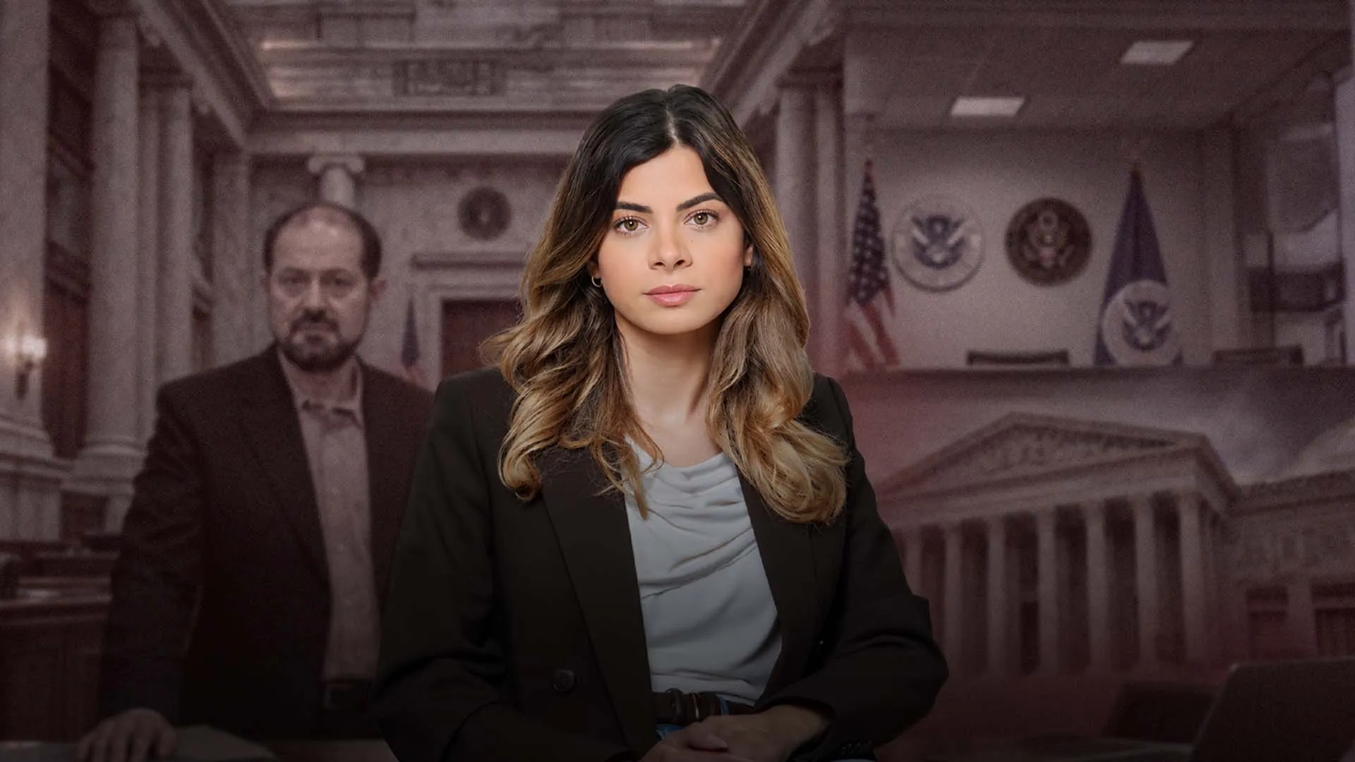 A woman in a black blazer sits before a blurred man and courtroom-style interior.