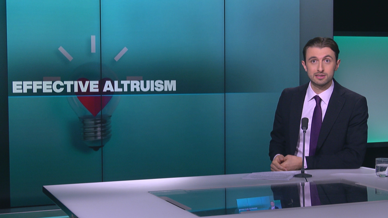 A suited presenter sits at a studio desk beside a screen displaying “Effective Altruism.”