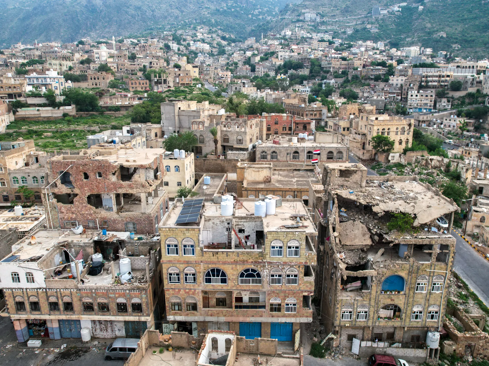 Dense hillside city with closely packed buildings, rooftop water tanks, solar panels, flags, and damaged structures.