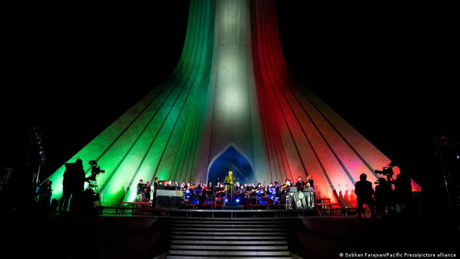 An orchestra performs beneath a monument illuminated in green, white, and red, with cameras filming from the sides.