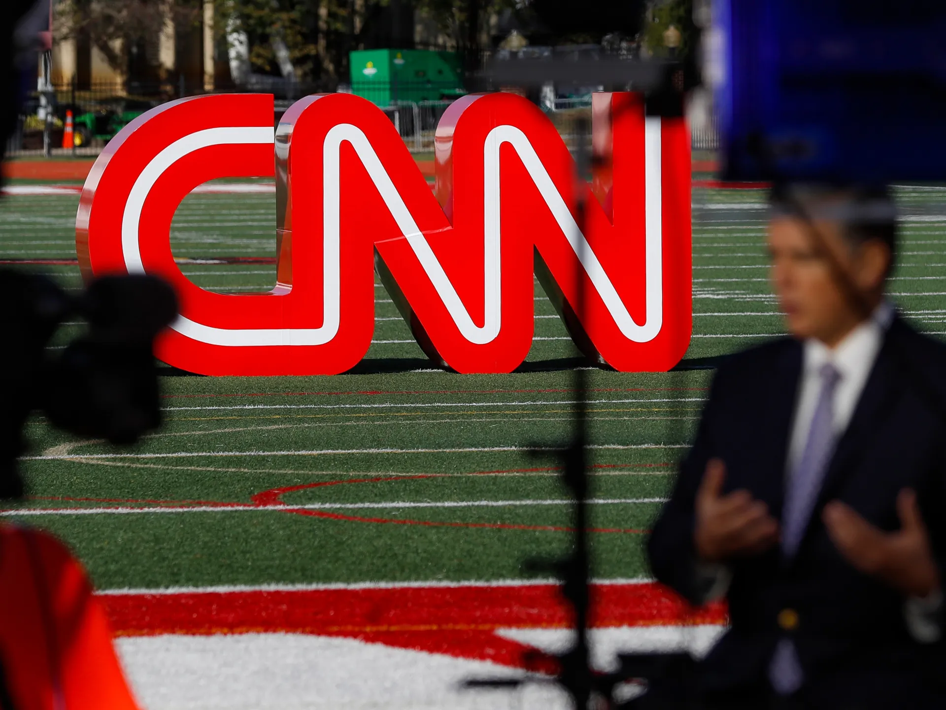 Large red CNN letters stand on a football field, with a blurred suited figure in the foreground.